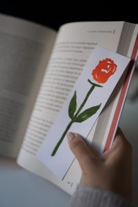 white bookmark with red rose and green stem on it held by a hand in an open book