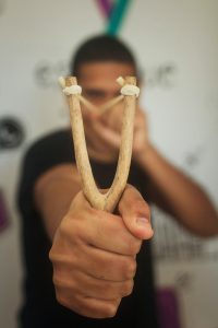 Man holding a slingshot and pointing it at the camera