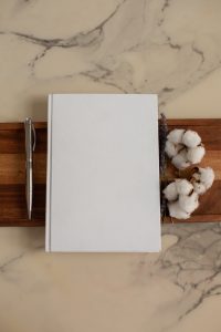 white marble background, brown wooden slab laying under a white journal, silver pin, and 3 bolls of cotton